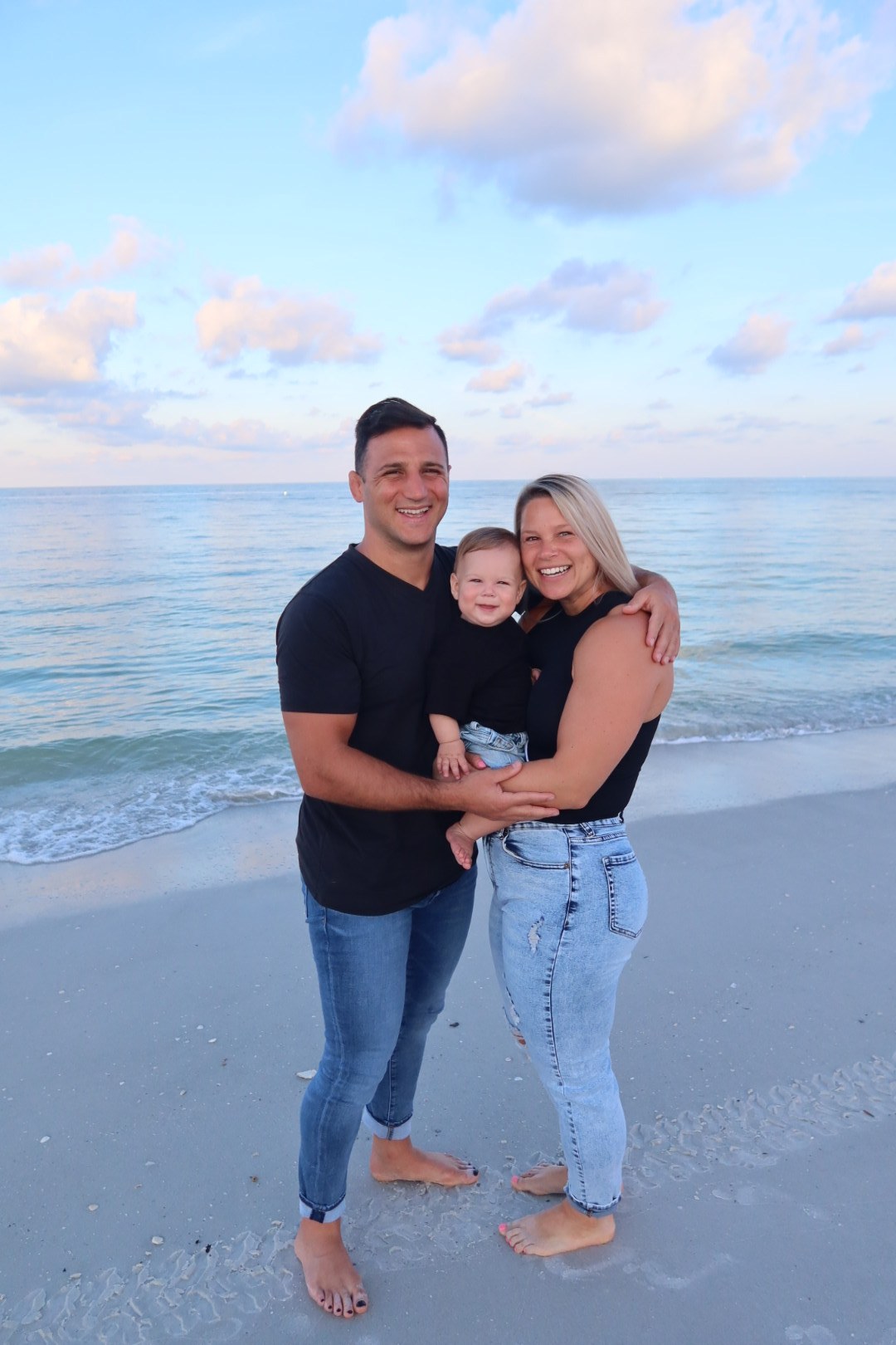 Joe, Carissa, and their son barefoot on the sand at dusk, the water calm behind them