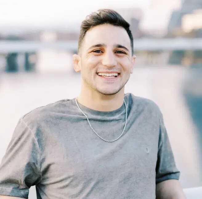 Joe Spisak smiling in a gray t-shirt, the Austin riverfront out of focus behind him