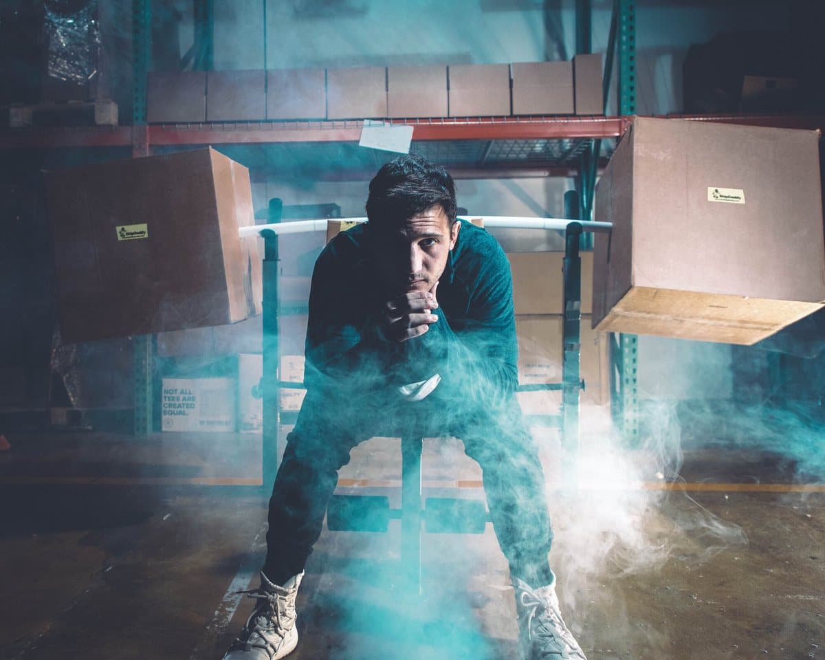 Joe crouched over a barbell loaded with ShipDaddy shipping boxes as plates, teal smoke drifting across the warehouse floor